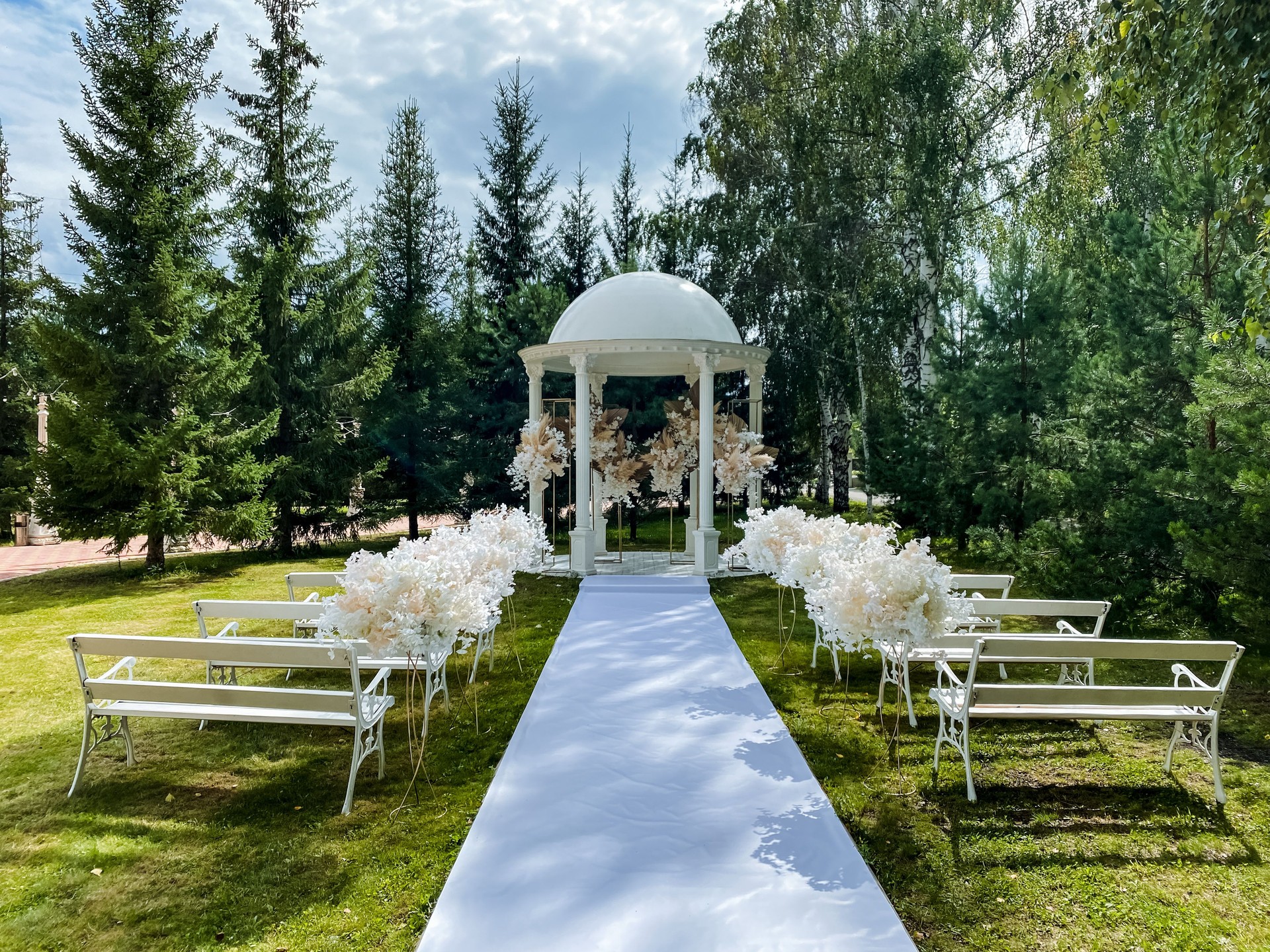 In a lovely park, a beautiful wedding ceremony is currently taking place, featuring a charming gazebo and inviting benches for guests to comfortably enjoy this wonderful and special event